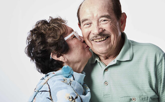 Picture of an elderly couple where the woman is kissing her husband on the cheek. 