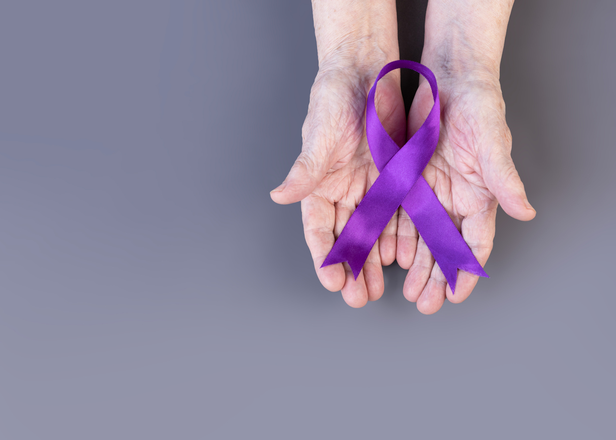 Hands of an elderly woman holding a purple ribbon for Alzheimer’s Awareness Month on a gray background