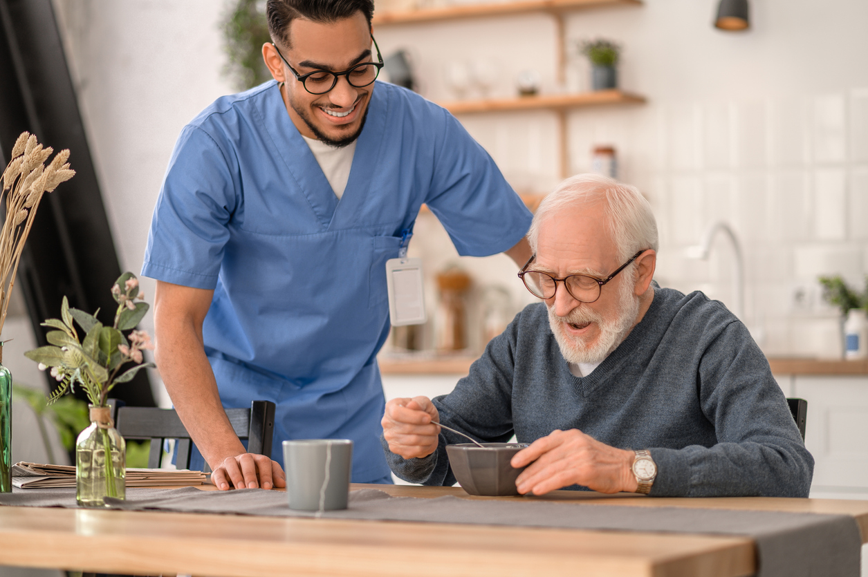 A caregiver with an older adult at a kitchen table in a home setting.