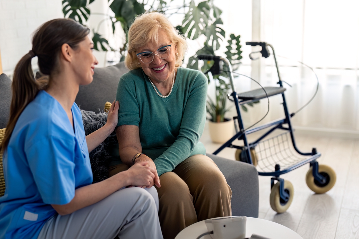 A woman sitting on a couch next to an older lady with a walker in the background.