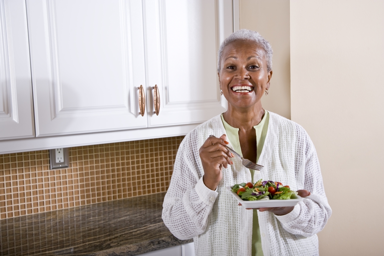 An older African American woman smiles at the camera while holding a plate of salad.