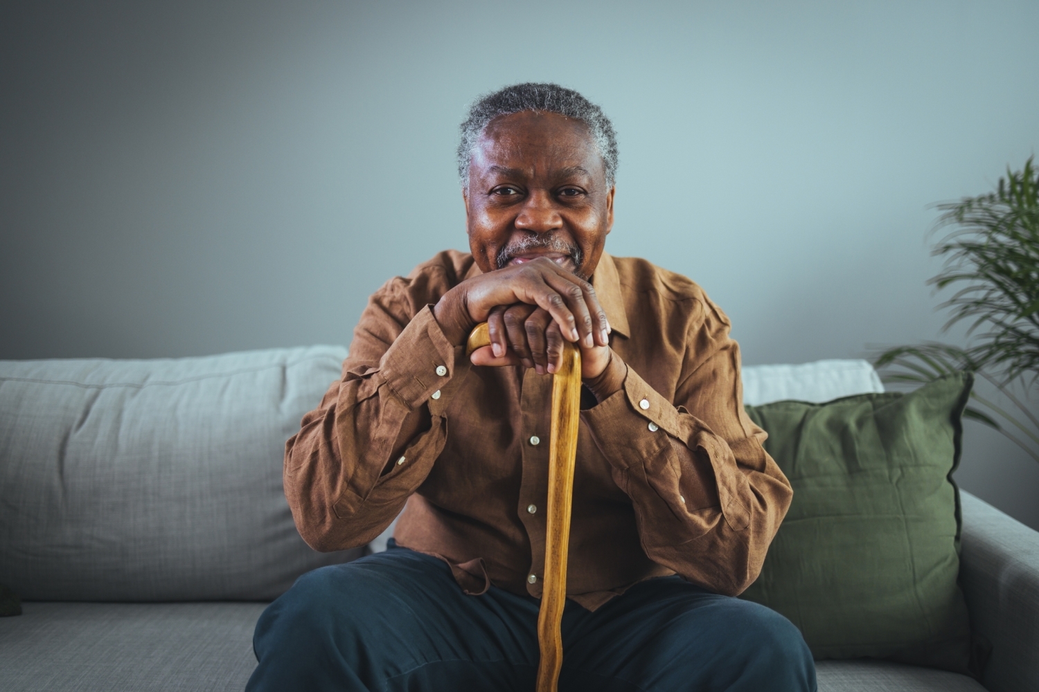 An older gentleman sitting on a couch leaning his head and hands on a cane, smiling.