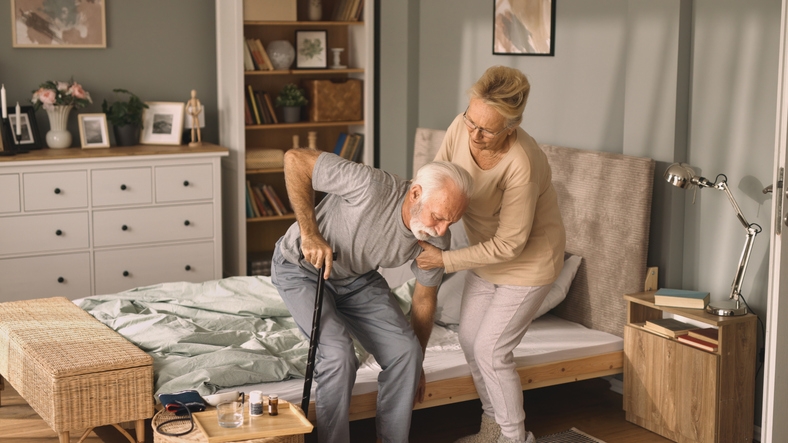 A lady helping an older gentleman get out of bed.
