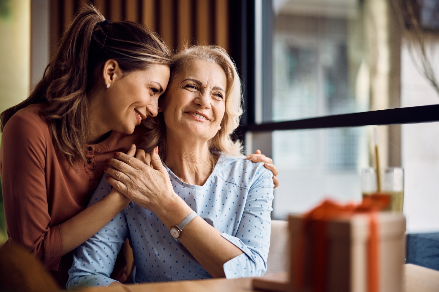 An adult daughter hugging her Mom while sitting at a table.