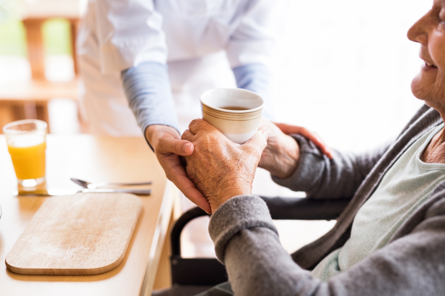 A caregiver helping to steady the hands of an older person to assist them in drinking their coffee.