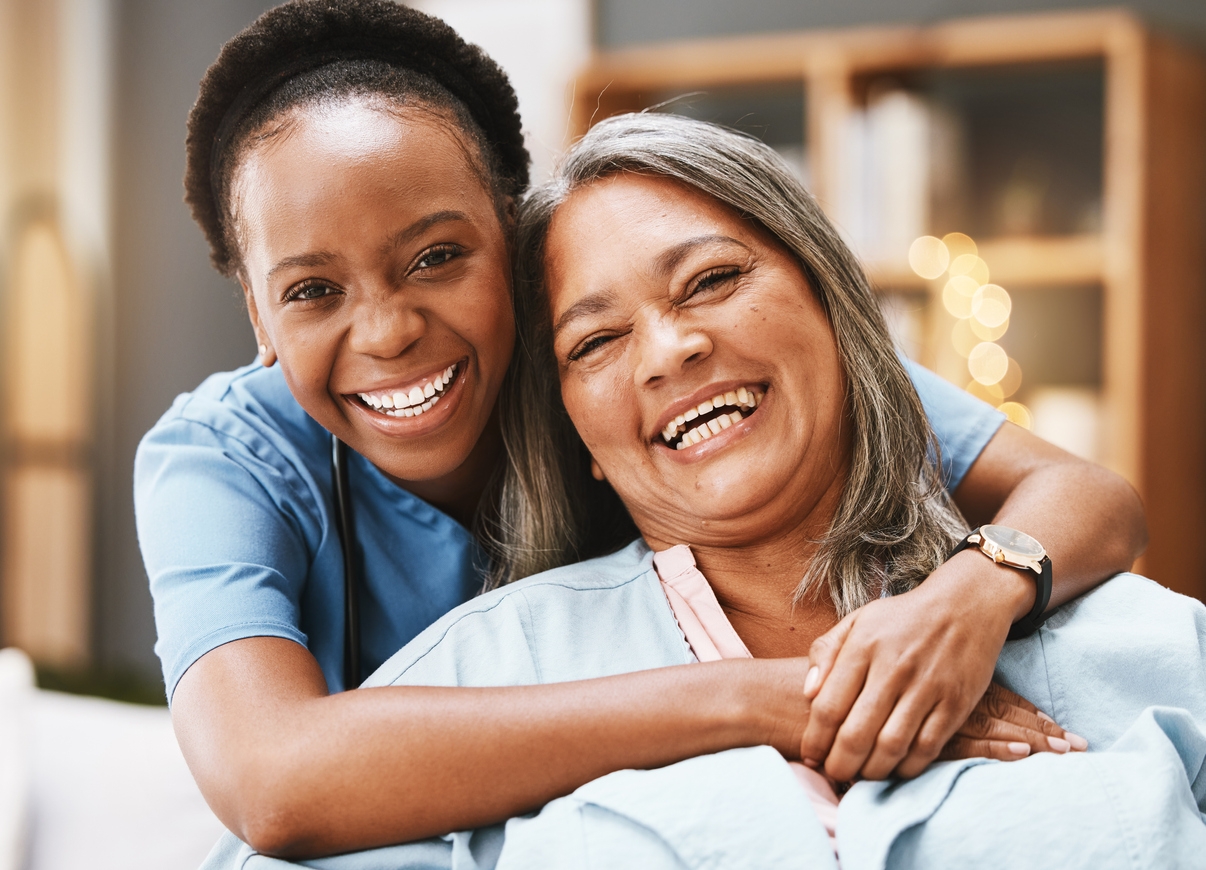 A caregiver happily hugging an older woman from behind as they pose for a photo.