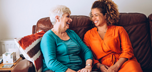 A caregiver sitting on a couch with an older woman laughing and talking.