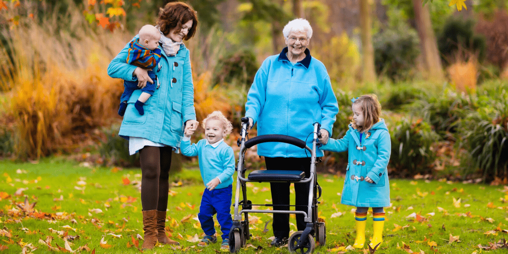 An older lady with a walker with a younger woman and 3 kids in the grass on a Fall day.