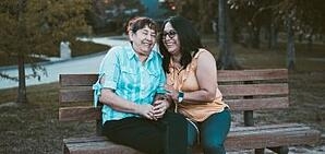 A pair of ladies sitting on a park bench talking and laughing.