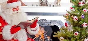 Santa Claus reading a storybook in front of a Christmas Tree.